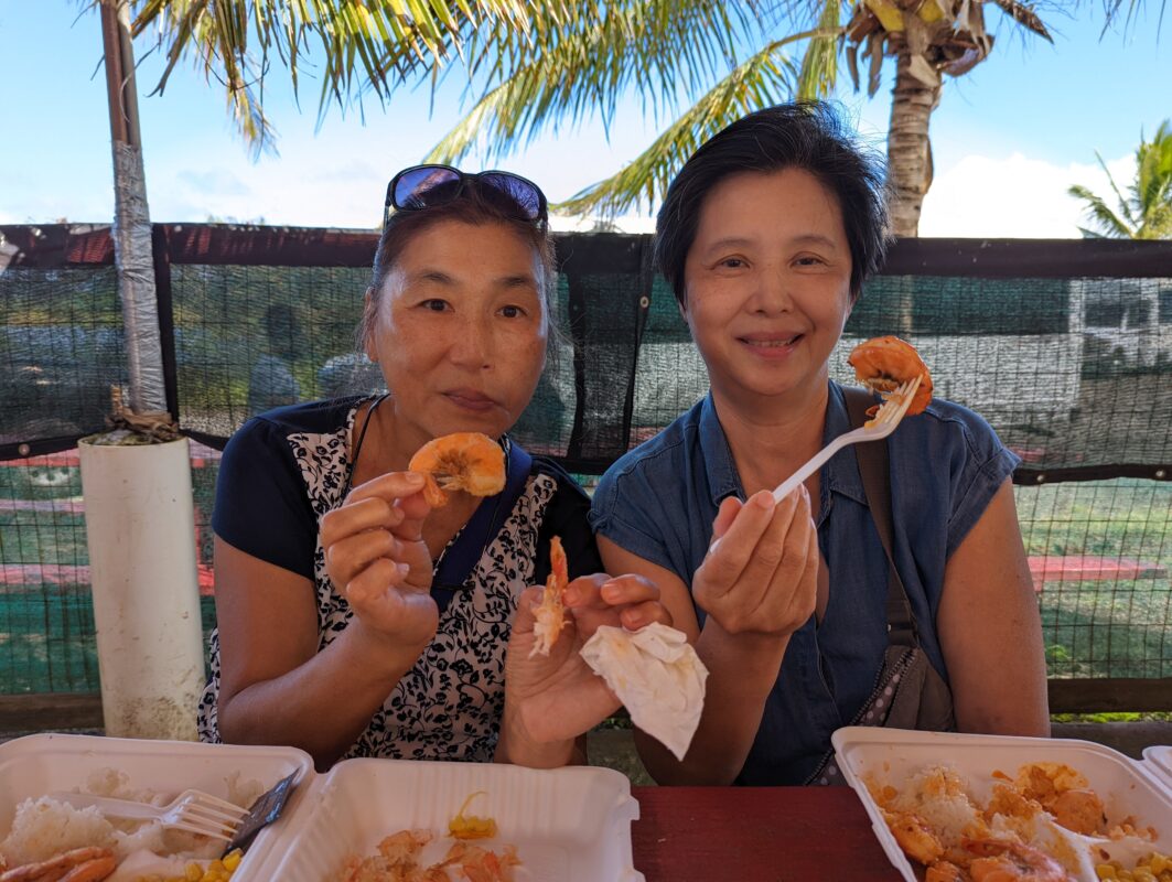 Two older women holding up shrimp and smiling.