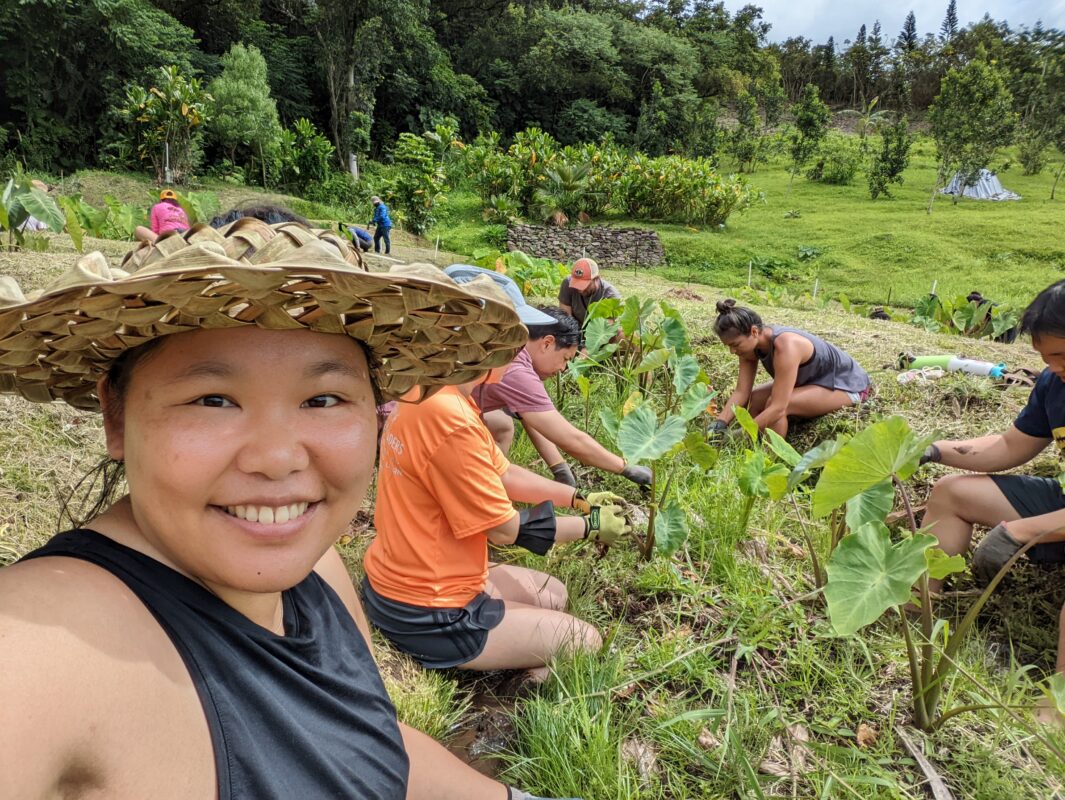 A woman smiling into the camera while people behind pull weeds.